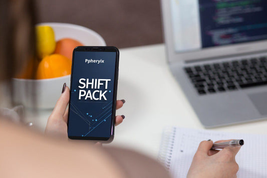 Person holding a phone with 'Ppheryix' app displaying 'Shift Pack' on a desk with a laptop and fruit bowl.